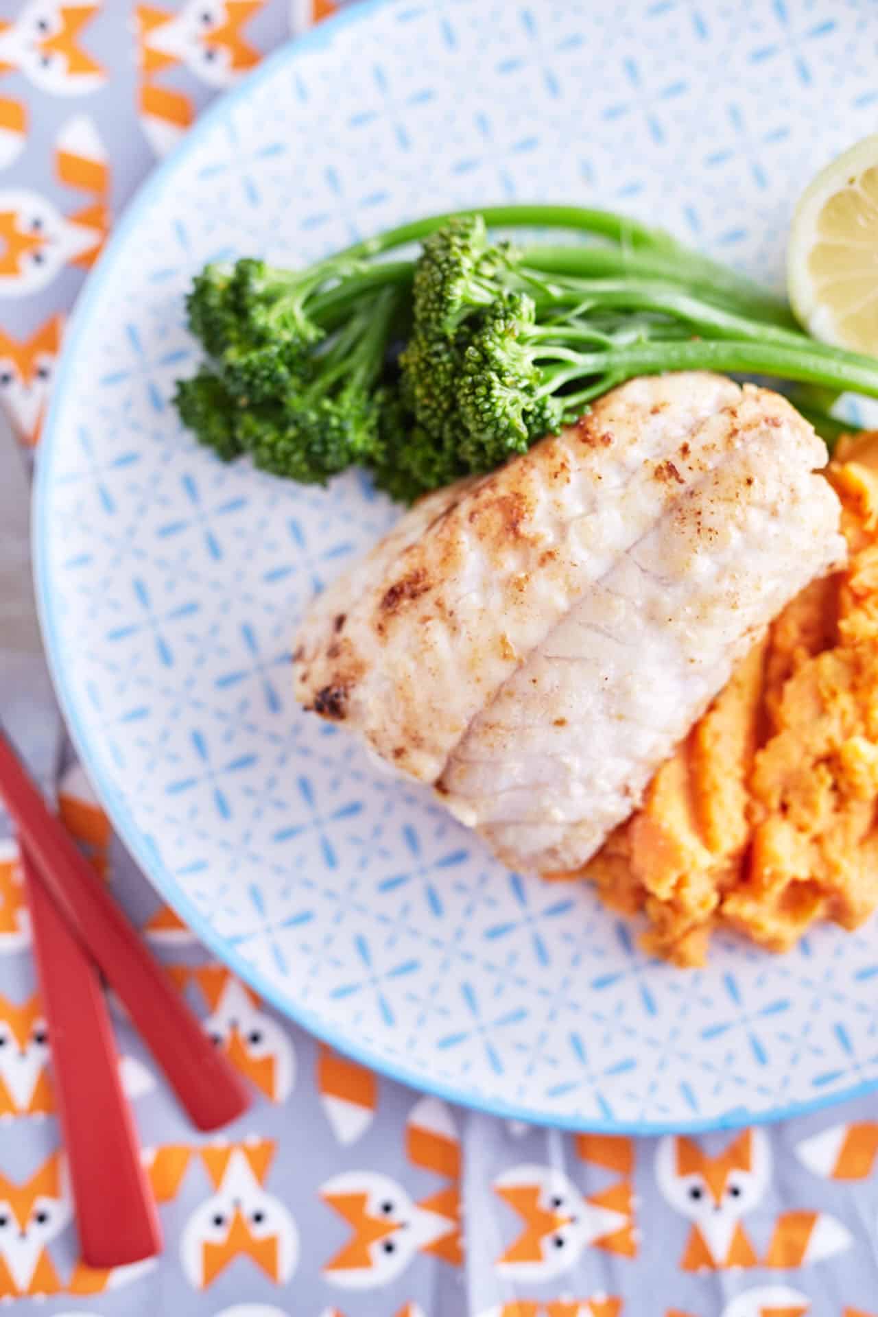 Pan fried white fish on mashed sweet potato with and broccolini with patterned plate on baby fox printed table cloth with red utensils