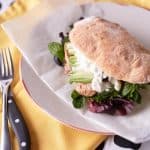 Jerk Chicken Sandwich with avocado and mesclun on a Turkish bun, dressed with sour cream sauce on a white plate with baking paper.