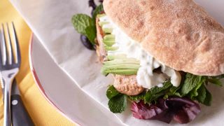 Jerk Chicken Sandwich with avocado and mesclun on a Turkish bun, dressed with sour cream sauce on a white plate with baking paper.