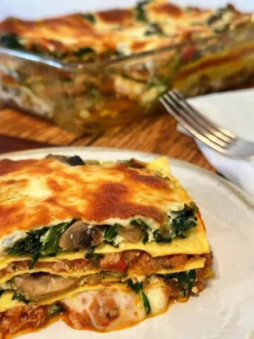 A slice of lasagna with layers of lasagna sheets, oozing with lamb sauce, melted cheese and spinach and a big serving dish with lamb lasagne in the background.