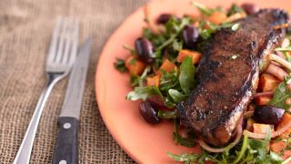 Juicy steak on bed of arugula salad with cubes of sweet potatoes, olives and red onion on a orange plate