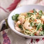 Plump, slightly pink-orange cooked shrimp atop angel hair pasta, garnished with fresh cilantro and chili, presented on a white plate.