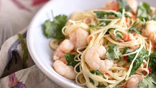 Plump, slightly pink-orange cooked shrimp atop angel hair pasta, garnished with fresh cilantro and chili, presented on a white plate.