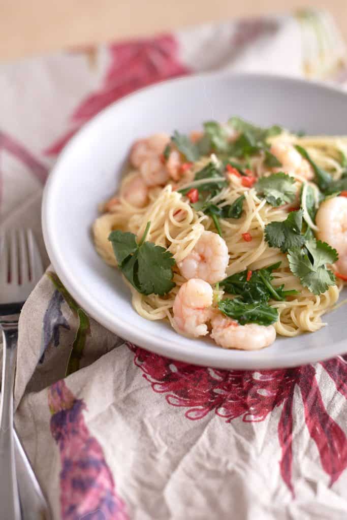 Plump, slightly pink-orange cooked shrimp atop angel hair pasta, garnished with fresh cilantro and chili, presented on a white plate.