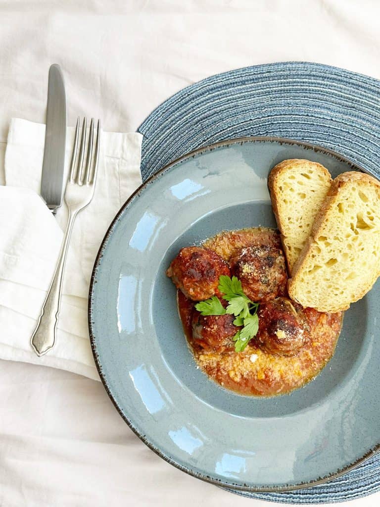 Four baked meatballs covered in marinara sauce and Parmesan, garnished with fresh parsley, presented on a blue plate alongside a side of crusty bread.