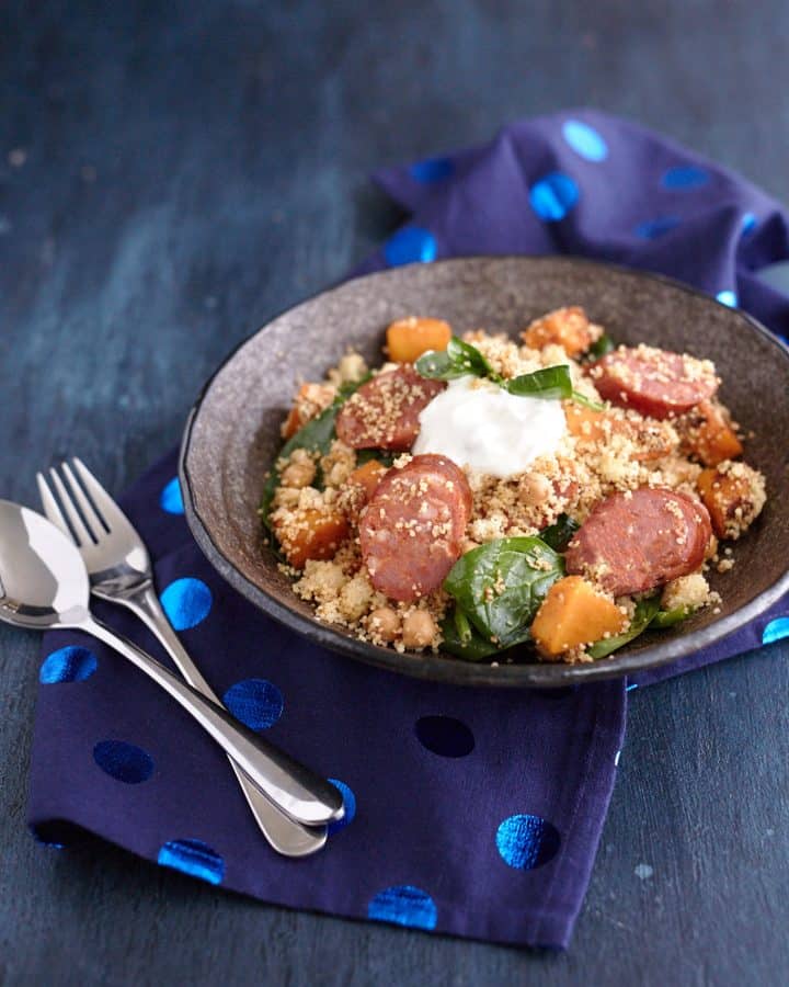 Sliced Chorizo sausage with cubed pumpkin, couscous, spinach, and chickpeas, garnished with a dollop of Greek yogurt served in a grey bowl.