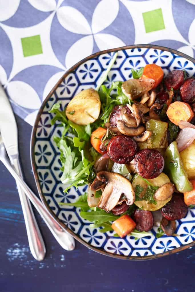 A Spanish chorizo recipe: A colourful array of chorizo and vegetables arranged on a bed of rocket leaves, presented on a patterned blue and white plate.