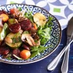 A colourful array of vegetables, accompanied by sliced and caramelised chorizo, arranged on a bed of rocket leaves, presented on a patterned blue and white plate.