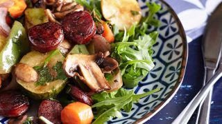 A colourful array of vegetables, accompanied by sliced and caramelised chorizo, arranged on a bed of rocket leaves, presented on a patterned blue and white plate.