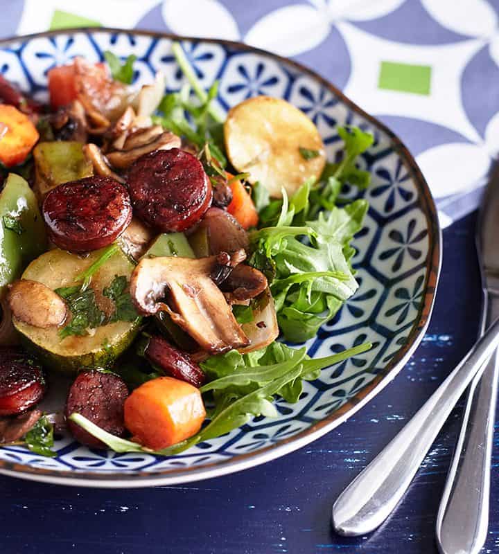 A colourful array of vegetables, accompanied by sliced and caramelised chorizo, arranged on a bed of rocket leaves, presented on a patterned blue and white plate.