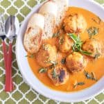 A white bowl holds a vibrant tomato and carrot soup with six neatly arranged meatballs, topped with fresh herbs, and served alongside crusty bread.