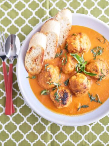 A white bowl holds a vibrant tomato and carrot soup with six neatly arranged meatballs, topped with fresh herbs, and served alongside crusty bread.