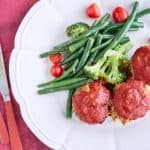 Three baby meatloaf muffins coated in vibrant red passata glaze are served on a white plate with steamed broccoli, green beans, and halved cherry tomatoes, set against a magenta background.