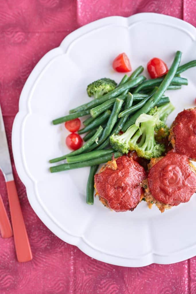 Three baby meatloaf muffins coated in vibrant red passata glaze are served on a white plate with steamed broccoli, green beans, and halved cherry tomatoes, set against a magenta background.