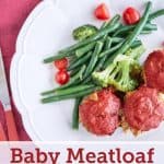 Three baby meatloaf muffins coated in vibrant red passata glaze are served on a white plate with steamed broccoli, green beans, and halved cherry tomatoes, set against a magenta background.