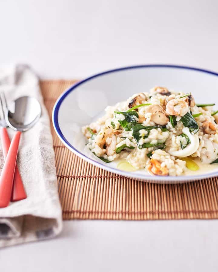 Seafood risotto features mussels, prawns, and calamari mixed with rice, served on a white plate with spinach leaves and a drizzle of olive oil.