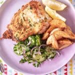 On a light pink plate atop a patterned tablecloth, you'll find golden brown fried fish lightly coated with specks of parsley, accompanied by golden roasted potato wedges and a vibrant green cucumber and red onion salad.