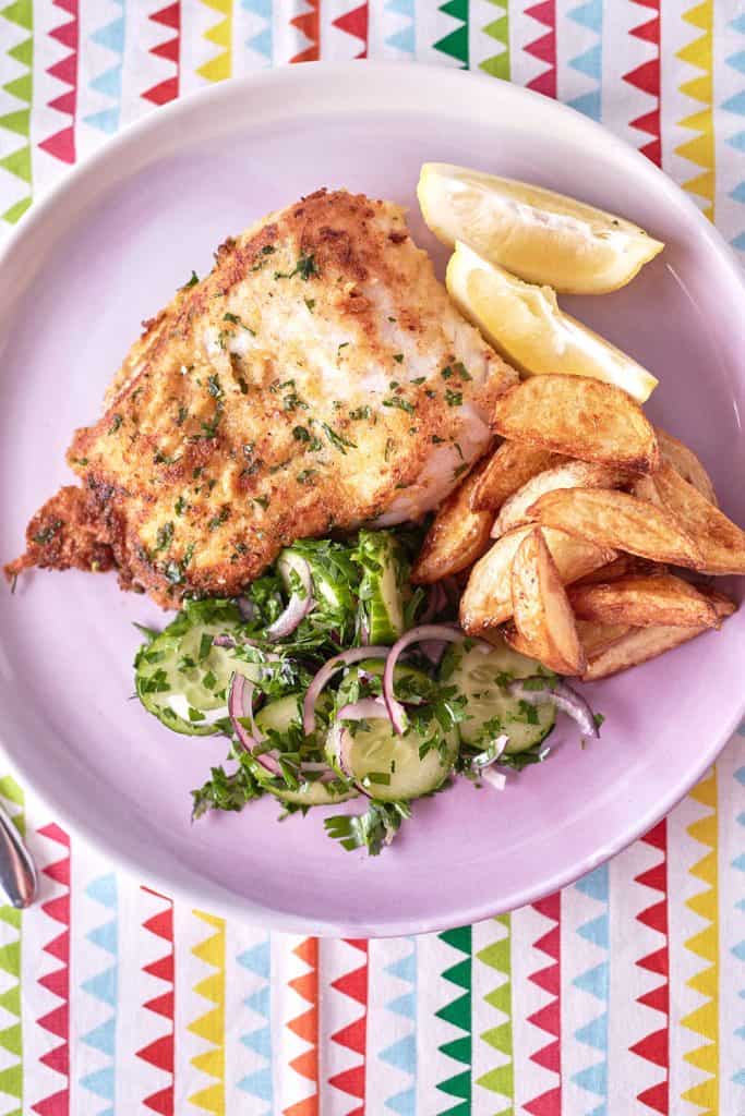 On a light pink plate atop a patterned tablecloth, you'll find golden brown fried fish lightly coated with specks of parsley, accompanied by golden roasted potato wedges and a vibrant green cucumber and red onion salad.