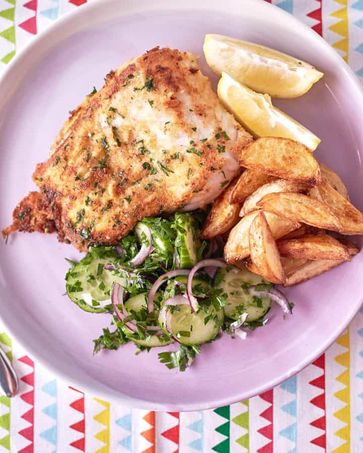 On a light pink plate atop a patterned tablecloth, you'll find golden brown fried fish lightly coated with specks of parsley, accompanied by golden roasted potato wedges and a vibrant green cucumber and red onion salad.