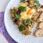 A patterned plate holds sliced baked potatoes with red onion and chorizo, topped by a cheesy baked egg and garnished with parsley, served alongside crusty bread.