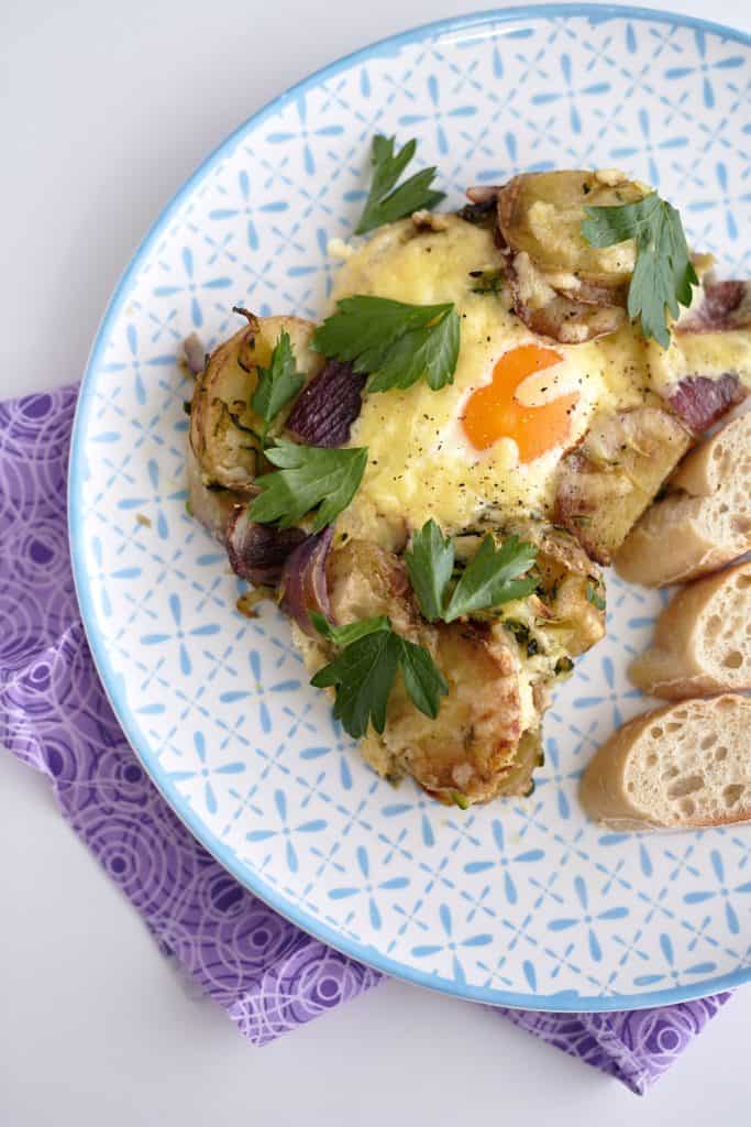 A patterned plate holds sliced baked potatoes with red onion and chorizo, topped by a cheesy baked egg and garnished with parsley, served alongside crusty bread.