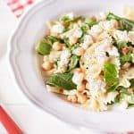 Farfalle pasta is mixed with chickpeas, fresh herbs, and specks of red chili, all coated in a lemon oil dressing. The dish is presented on a white plate.