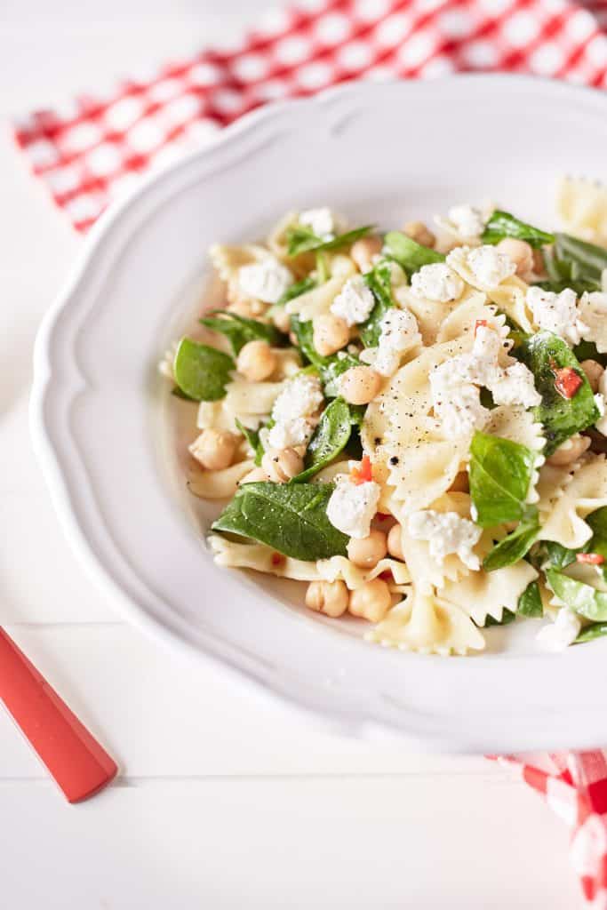 Farfalle pasta is mixed with chickpeas, fresh herbs, and specks of red chili, all coated in a lemon oil dressing. The dish is presented on a white plate.