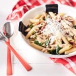 Mushroom and sun-dried tomato pasta with chicken, garnished with grated Parmesan cheese, is presented on a white plate atop a white table draped with a red and white chequered tablecloth.