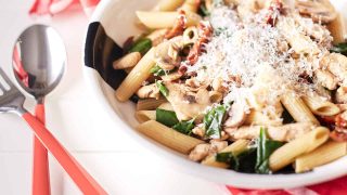 Mushroom and sun-dried tomato pasta with chicken, garnished with grated Parmesan cheese, is presented on a white plate atop a white table draped with a red and white chequered tablecloth.