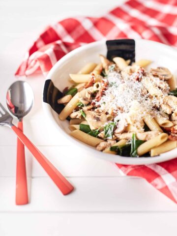 Mushroom and sun-dried tomato pasta with chicken, garnished with grated Parmesan cheese, is presented on a white plate atop a white table draped with a red and white chequered tablecloth.