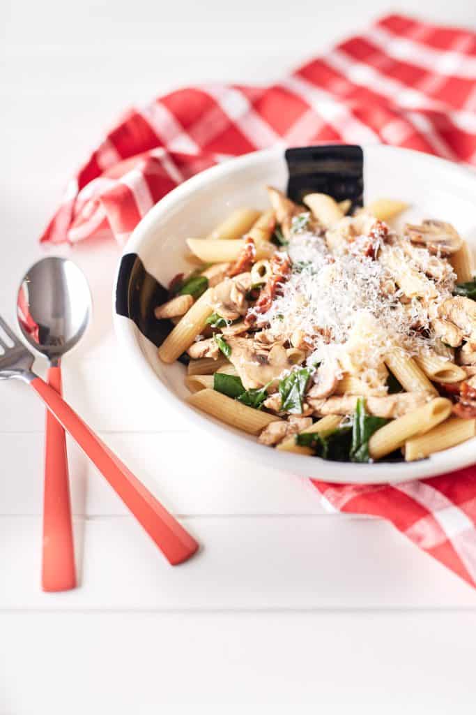 Mushroom and sun-dried tomato pasta with chicken, garnished with grated Parmesan cheese, is presented on a white plate atop a white table draped with a red and white chequered tablecloth.