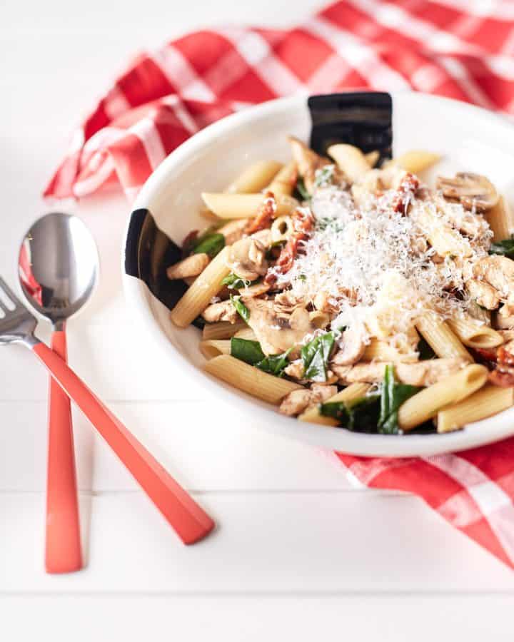 Mushroom and sun-dried tomato pasta with chicken, garnished with grated Parmesan cheese, is presented on a white plate atop a white table draped with a red and white chequered tablecloth.