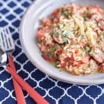 Tomato risotto with sliced sausages topped with basil and parmesan on a grey plate, placed on a blue and white tablecloth.