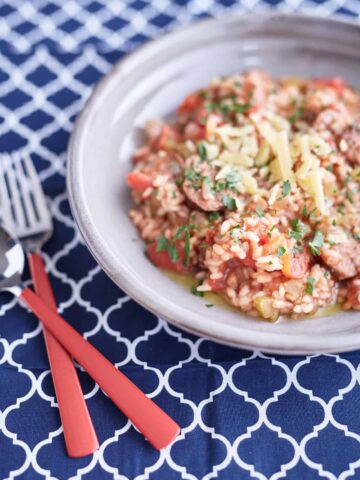 Tomato risotto with sliced sausages topped with basil and parmesan on a grey plate, placed on a blue and white tablecloth.