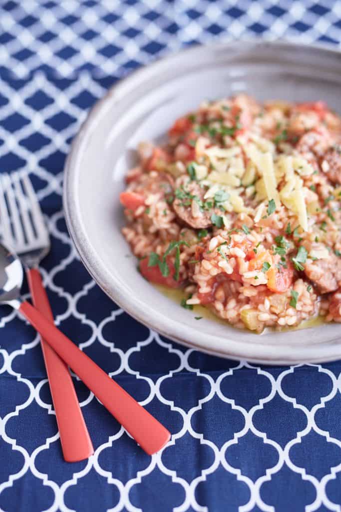 Tomato risotto with sliced sausages topped with basil and parmesan on a grey plate, placed on a blue and white tablecloth.