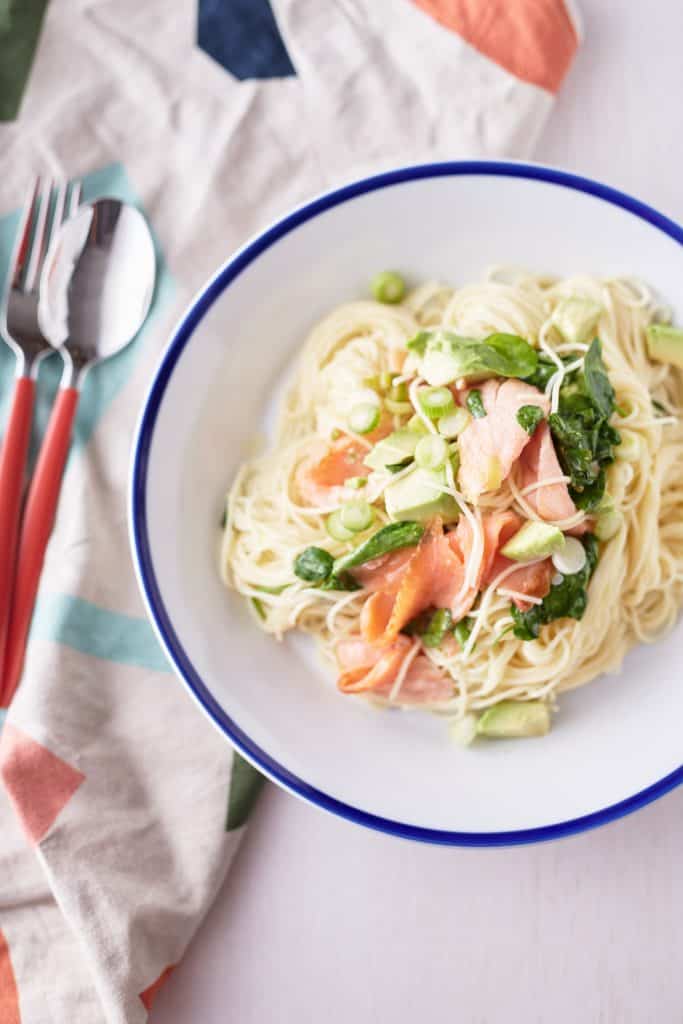 White plate with delicate angel hair pasta topped with slices of smoked salmon, cubed avocado, fresh watercress, and thinly sliced spring onions.