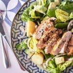 Golden brown pan-fried sliced chicken breast rests atop a bed of crisp cos lettuce, accompanied by halved baby potatoes, cubed avocado, red onion, and snow peas, arranged on a blue patterned plate.