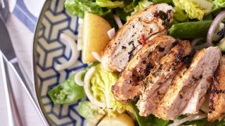 Golden brown pan-fried sliced chicken breast rests atop a bed of crisp cos lettuce, accompanied by halved baby potatoes, cubed avocado, red onion, and snow peas, arranged on a blue patterned plate.