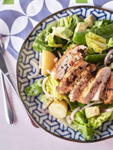 Golden brown pan-fried sliced chicken breast rests atop a bed of crisp cos lettuce, accompanied by halved baby potatoes, cubed avocado, red onion, and snow peas, arranged on a blue patterned plate.