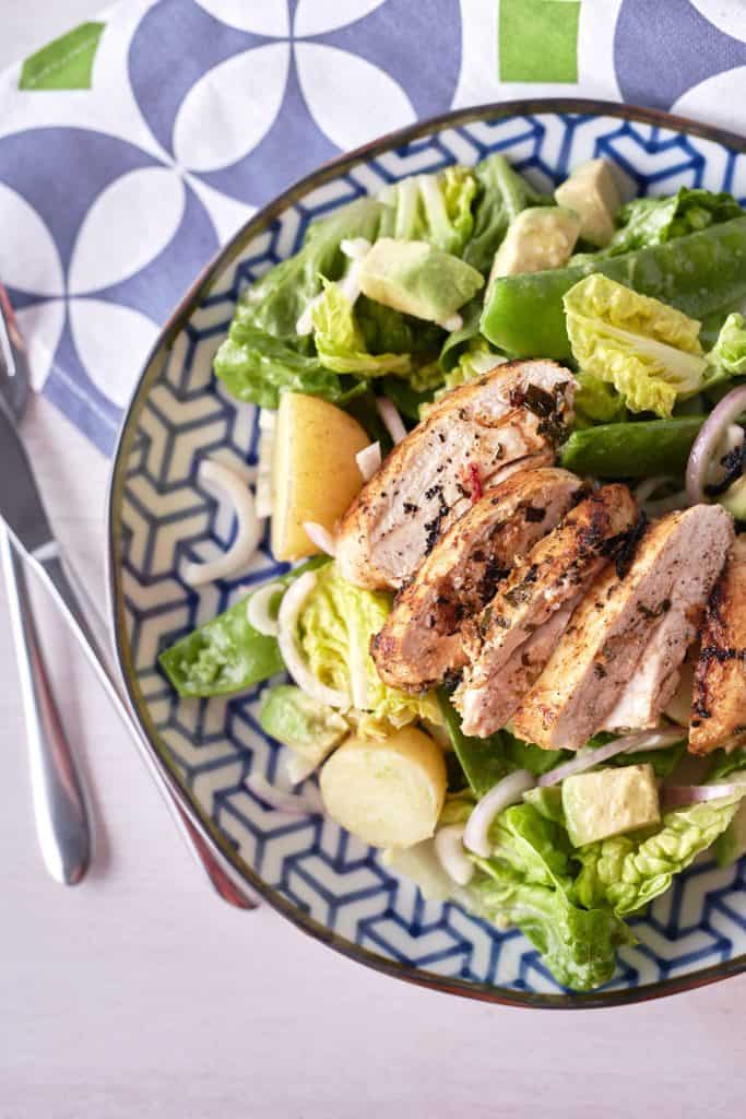 Spicy chicken salad - Golden brown pan-fried sliced chicken breast rests atop a bed of crisp cos lettuce, accompanied by halved baby potatoes, cubed avocado, red onion, and snow peas, arranged on a blue patterned plate.