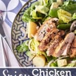 Golden brown pan-fried sliced chicken breast rests atop a bed of crisp cos lettuce, accompanied by halved baby potatoes, cubed avocado, red onion, and snow peas, arranged on a blue patterned plate.