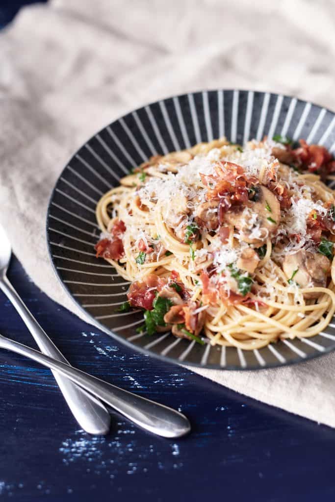 Crispy Prosciutto Pasta - Spaghetti, topped with crunchy prosciutto and sautéed mushrooms, garnished with parsley and Parmesan cheese, served on a black plate.