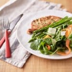 Green mixed lettuce leaves are intertwined with carrot ribbons, accompanied by asparagus spears, and a crispy golden-brown crumbed eggplant slice, all presented on a white plate.