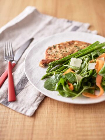 Green mixed lettuce leaves are intertwined with carrot ribbons, accompanied by asparagus spears, and a crispy golden-brown crumbed eggplant slice, all presented on a white plate.