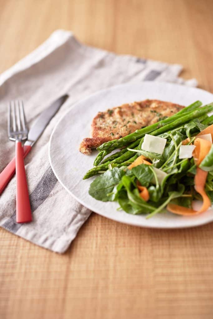 Green mixed lettuce leaves are intertwined with carrot ribbons, accompanied by asparagus spears, and a crispy golden-brown crumbed eggplant schnitzel slice, all presented on a white plate.