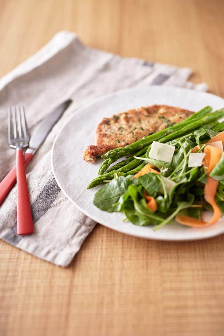 Green mixed lettuce leaves are intertwined with carrot ribbons, accompanied by asparagus spears, and a crispy golden-brown crumbed eggplant slice, all presented on a white plate.