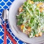 A creamy sage risotto with cubed pumpkin and snow peas, topped with rocket leaves, Parmesan cheese, and lemon zest, served on a light grey plate on a patterned blue tablecloth.