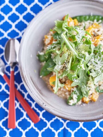 A creamy sage risotto with cubed pumpkin and snow peas, topped with rocket leaves, Parmesan cheese, and lemon zest, served on a light grey plate on a patterned blue tablecloth.