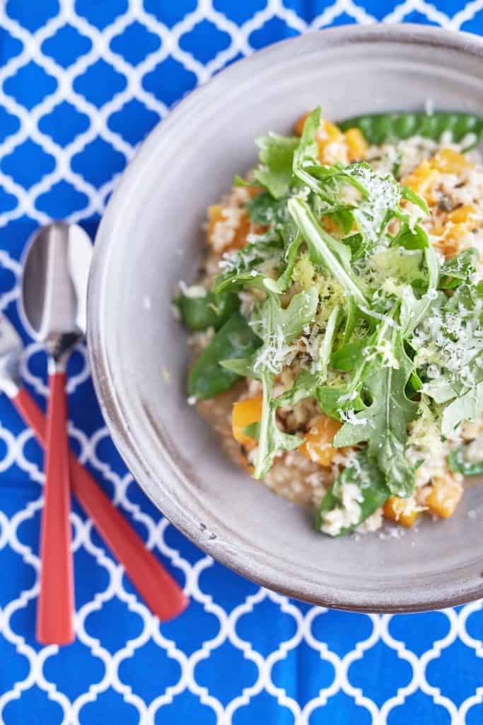 A creamy sage risotto with cubed pumpkin and snow peas, topped with rocket leaves, Parmesan cheese, and lemon zest, served on a light grey plate on a patterned blue tablecloth.