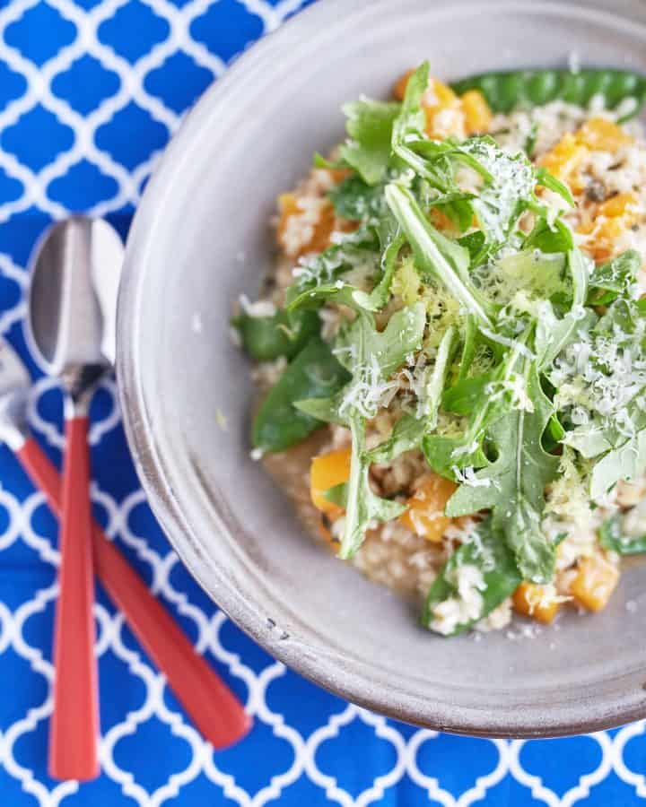 A creamy sage risotto with cubed pumpkin and snow peas, topped with rocket leaves, Parmesan cheese, and lemon zest, served on a light grey plate on a patterned blue tablecloth.